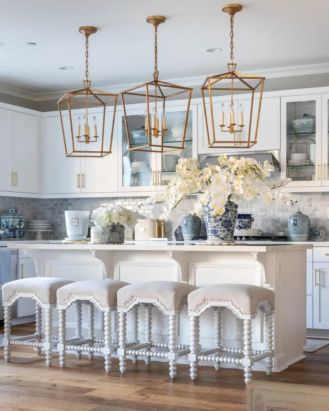 Bright, modern kitchen with white cabinets, gold pendant lights, a marble island with four cushioned stools, and blue and white vases—perfect inspiration for how to avoid common mistakes when decorating for seasonal changes.