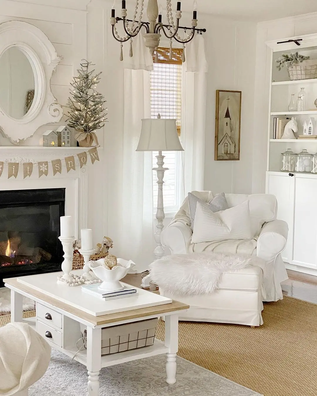 A cozy living room with white furniture, a fireplace, a decorated mantel, a small Christmas tree, and shelves showing neutral-toned decor—perfect inspiration for how to avoid common mistakes when decorating for seasonal changes.