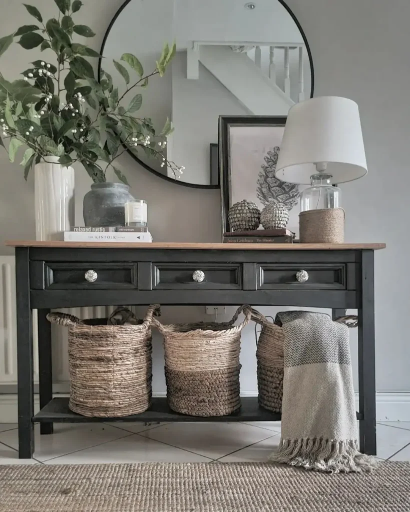 How to Design a Summer-Ready Entryway: A black console table with wicker baskets underneath, a lamp, books, vases with greenery, and framed art on top sits in front of a round mirror on a gray wall.