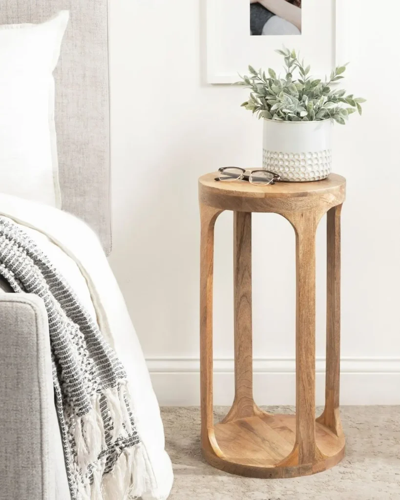 A wooden side table next to a bed holds a potted plant and eyeglasses, showcasing how to transform small spaces with minimalist side tables; a blanket is draped over the bed.