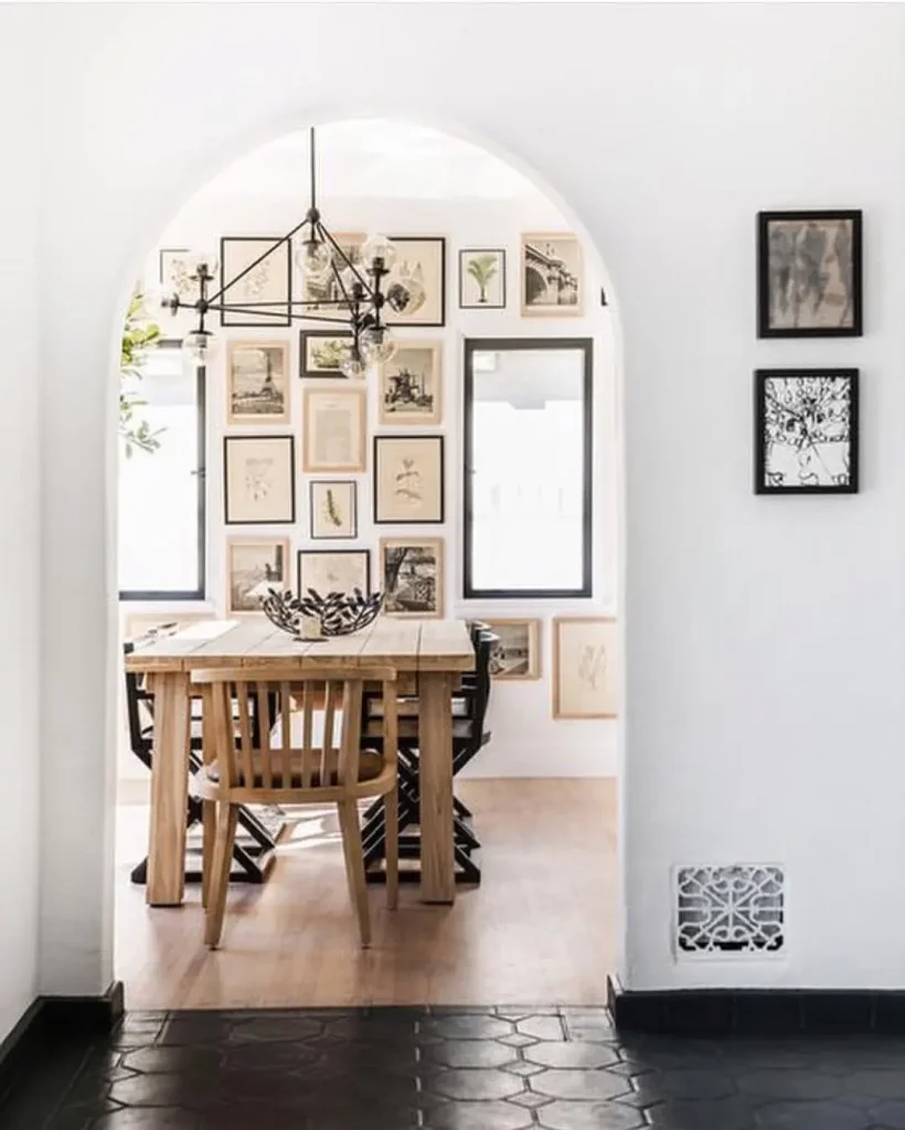 A wooden dining table with chairs sits under a black chandelier in a bright room, viewed through an arched doorway; the far wall showcases framed artwork, demonstrating how to create a stunning gallery wall for every room.