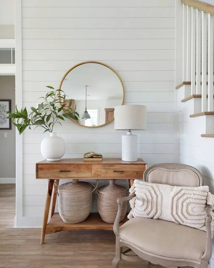 A wooden console table with two round baskets underneath, a round mirror, a lamp, and a vase with greenery. To the right, there's a beige cushioned armchair with a decorative pillow—perfect elements for how to design a summer-ready entryway.