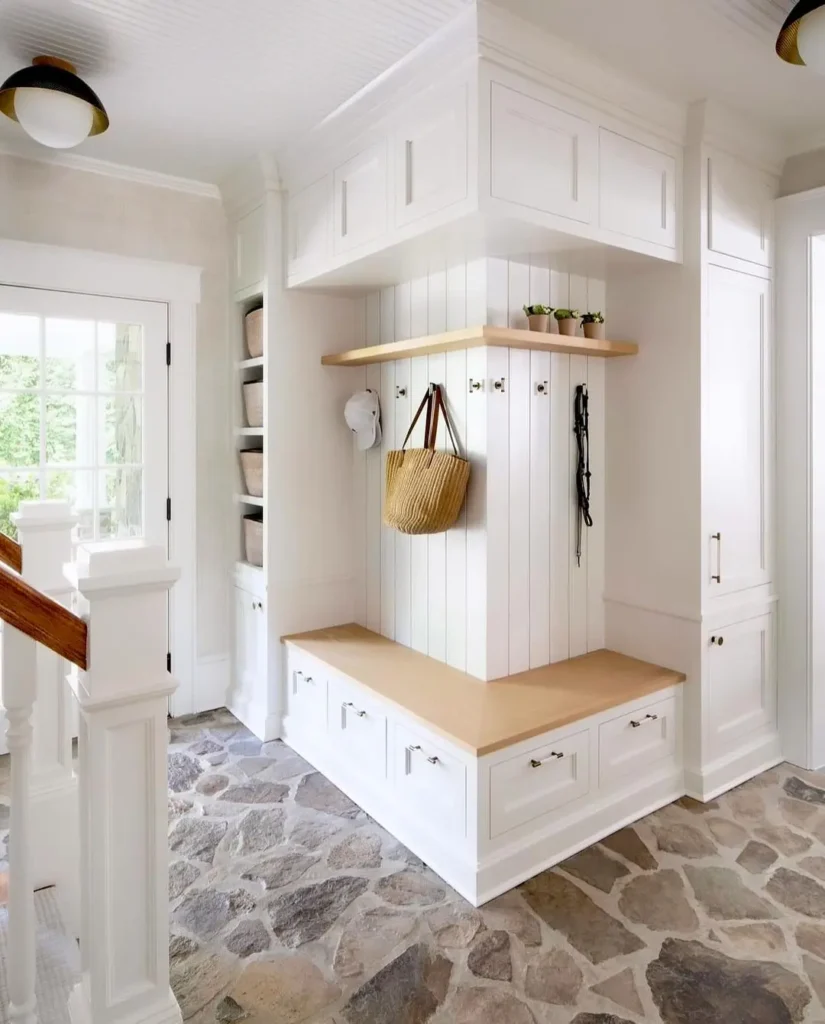 A bright entryway with stone flooring, white built-in cabinets, a wooden bench, hooks with a bag and leashes, small plants, and shelves with baskets—your ultimate functional and stylish mudroom guide in action.
