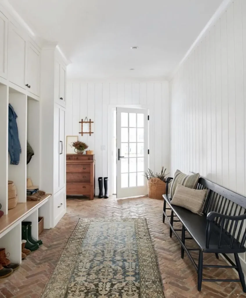 A bright, narrow mudroom with white paneled walls, built-in storage, a wooden bench, and a patterned rug showcases ideas from Your Ultimate Functional and Stylish Mudroom Guide, all illuminated by a glass door leading outside.