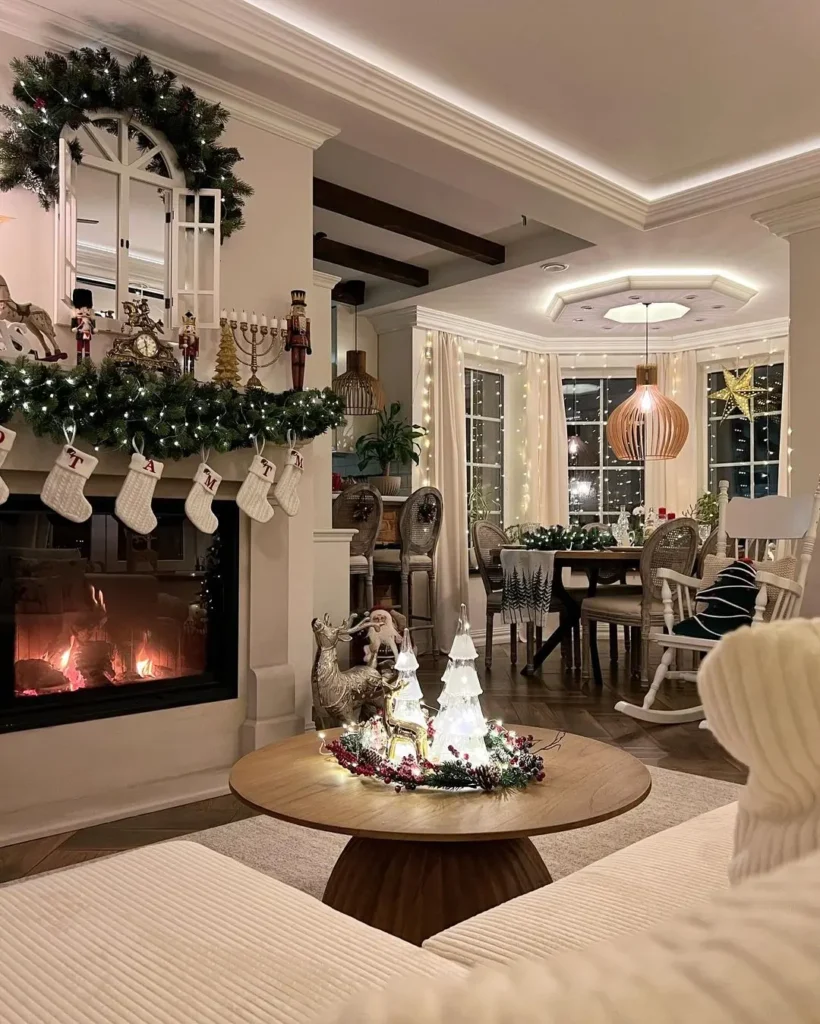 A cozy living room decorated for Christmas with stockings on the fireplace, a lit fire, festive ornaments, and a dining table set near large windows—one of 10 Cozy Christmas Living Room Ideas You’ll Love This Year.