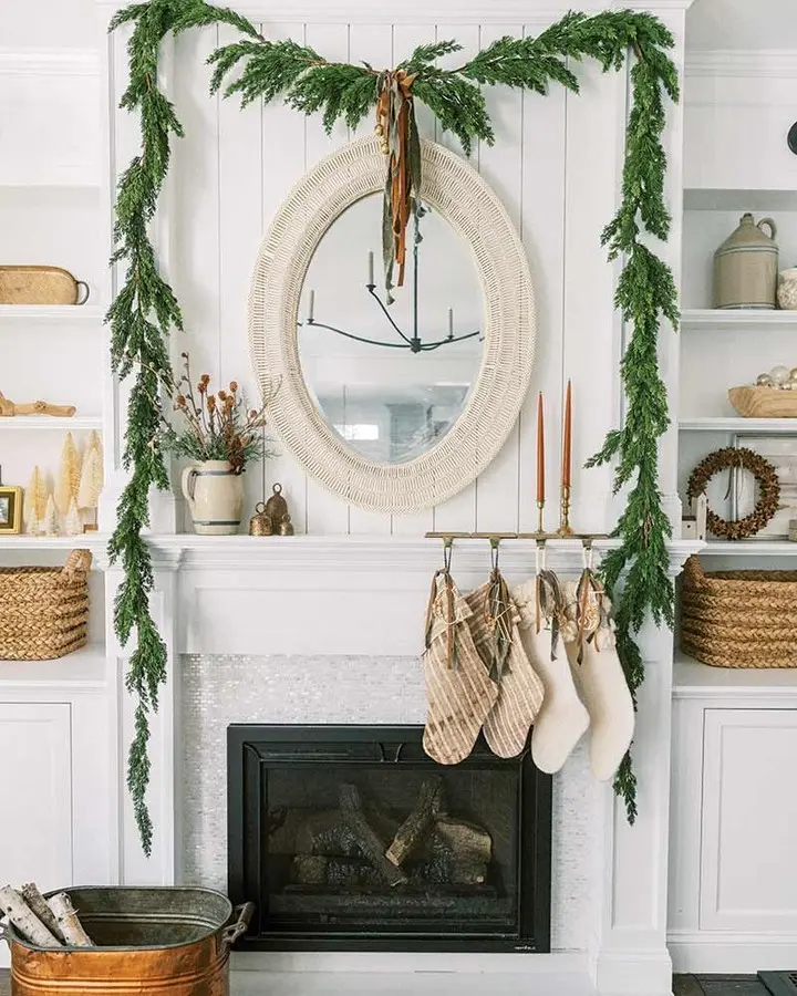 A white fireplace decorated with green garland, a round mirror, four stockings, and tall candles perfectly captures the spirit of 10 Stunning Christmas Mantel Ideas to Beautify Your Home, with neutral-toned holiday decor on surrounding shelves.