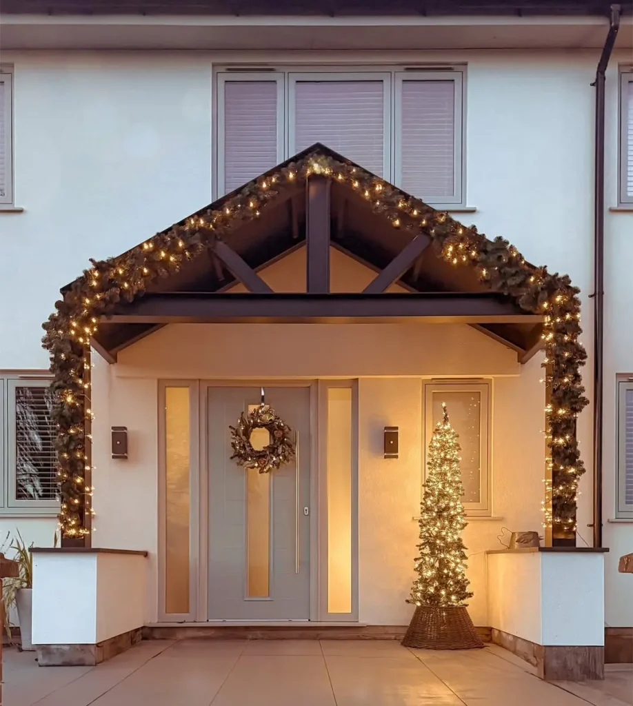 House entrance decorated with string lights—showing how to use string lights on your front porch—a wreath on the door, and a lit Christmas tree-shaped decoration by the doorway.