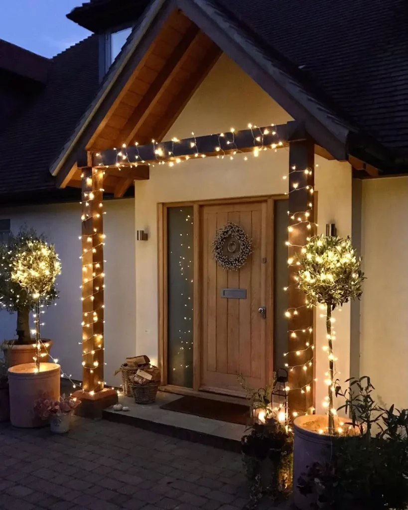 A house entrance with a wooden door is decorated with string lights wrapped around two columns and potted trees, offering inspiration on how to use string lights on your front porch for a warm, inviting atmosphere at dusk.