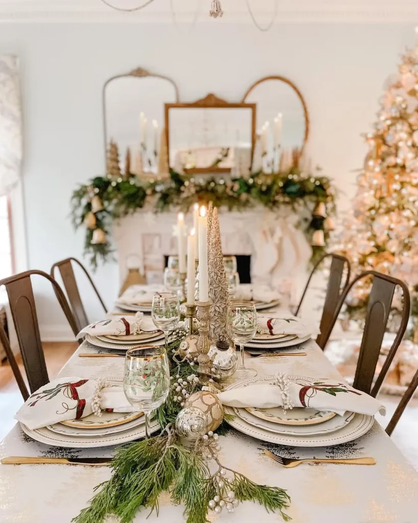 A dining table set for a holiday meal with festive decorations, candles, glassware, and greenery showcases easy Christmas table settings and centerpieces for any style, with a decorated Christmas tree and mantelpiece in the background.