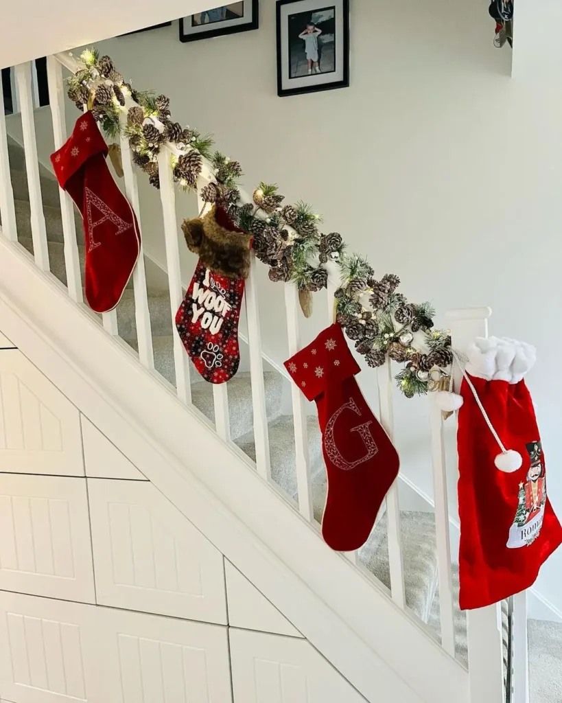 A staircase railing decorated with Christmas stockings, pinecones, garland, and lights—an idea inspired by 10 Adorable Kid-Friendly Christmas Decorations You’ll Love—with framed photos hanging on the wall in the background.