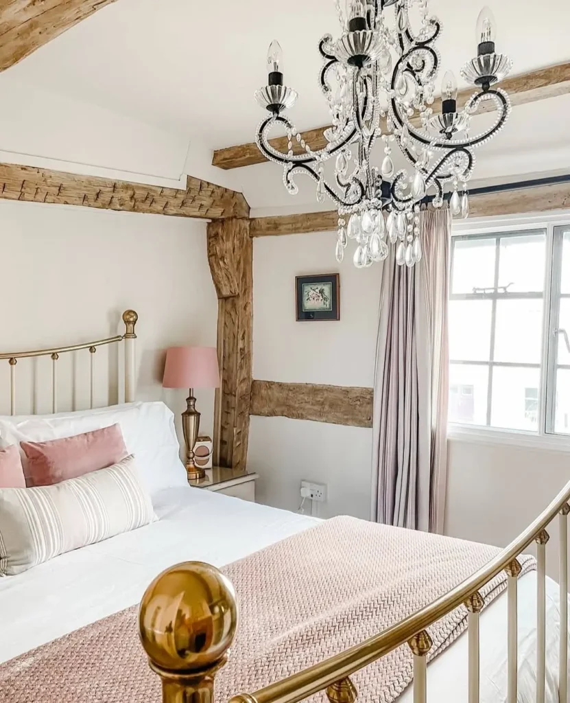 An affordable rustic bedroom featuring a brass bed, white and pink bedding, exposed wooden beams, a crystal chandelier, light curtains on the window, and a small framed picture on the wall.