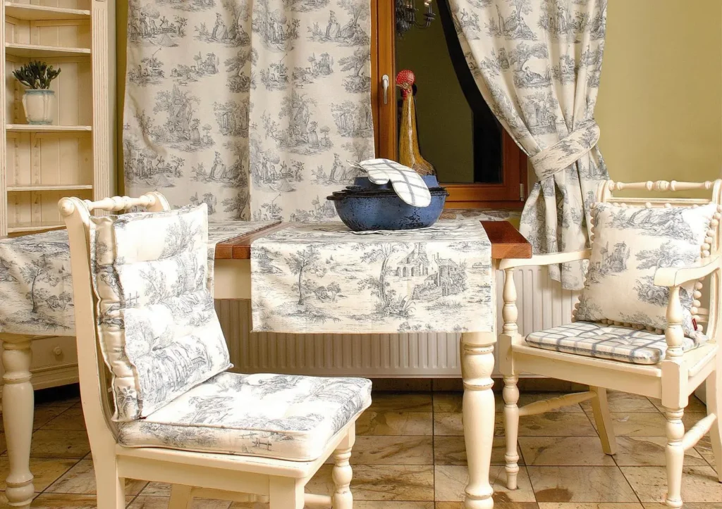 A cozy dining area with two chairs and a table, all featuring matching French country toile de Jouy pattern fabric. A ceramic chicken dish sits on the table, complemented by matching curtains in the background.
