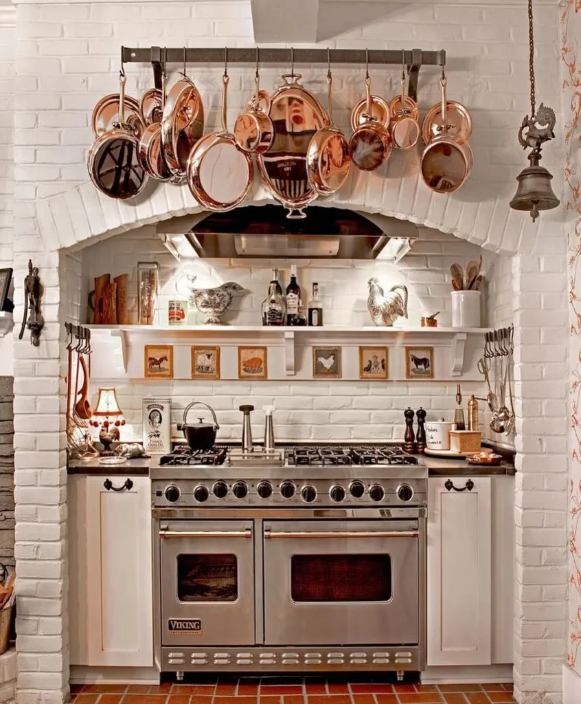 A kitchen with a french country vibe features a modern stove by Viking, white brick walls, shelves with decor, and copper pots and pans hanging above the stove.