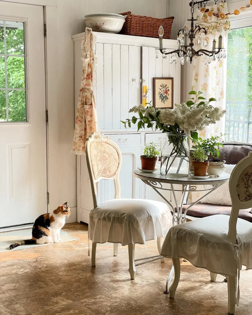 A calico cat sits by a white door in a bright, vintage-style kitchen with french country neutral paint color, white furniture, potted plants, and a glass table holding a vase of white flowers.