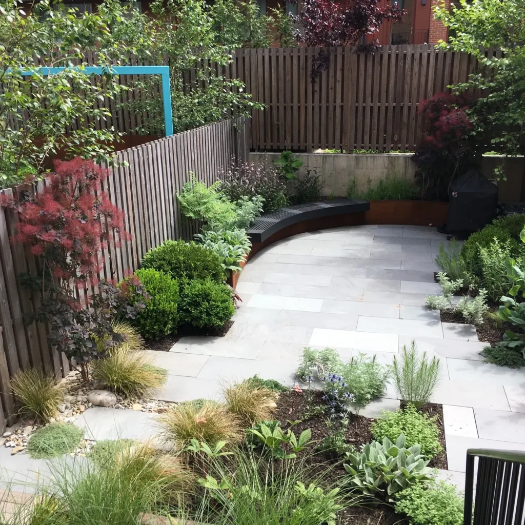 A landscaped backyard with stone paving, curved wooden bench, wooden fence, and lush green shrubs, grasses, and small trees creates a serene organic modern retreat.