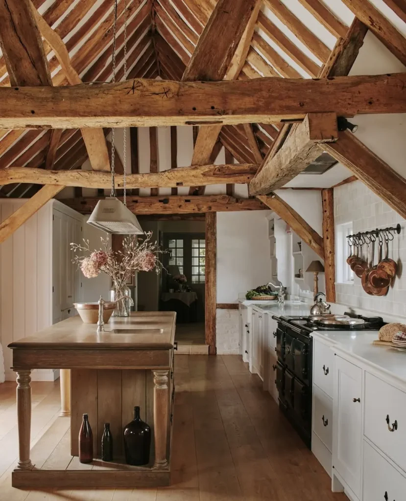 A rustic kitchen with exposed wooden beams, a wooden island with vases, white cabinets, a black stove, and hanging utensils on a white tile wall, perfect for inspiring your rustic kitchen ideas.