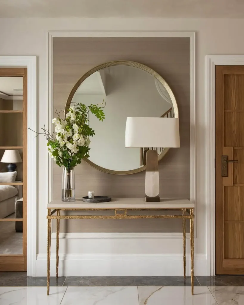 A modern console table with a gold frame stands against the wall, topped with a vase of white flowers and a lamp, beneath a large round mirror—an inviting touch of minimalist organic modern entryway decor between two wooden doors.