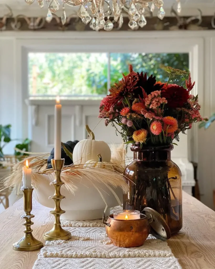A rustic fall dining table centerpiece with a wooden table, two brass candlesticks, a lit candle in a copper holder, a vase of autumn flowers, a white bowl with a white pumpkin, and wheat stalks.