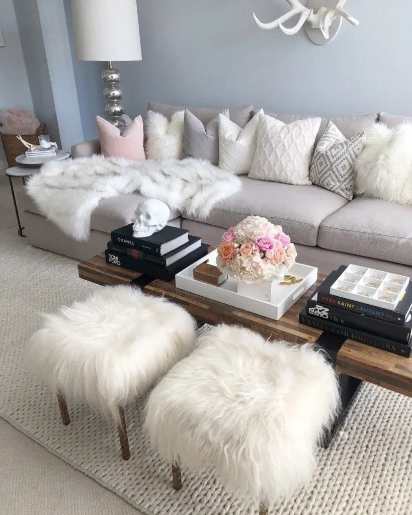 A modern living room styled for winter with a light gray sofa, white fur throws, fluffy stools, stacked books, a floral arrangement, and decorative accents on a coffee table.