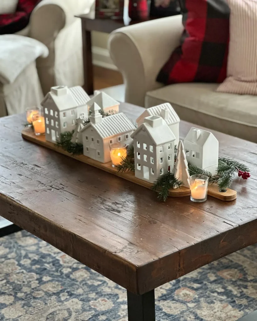 A wooden coffee table displays a miniature ceramic village with small candles, faux greenery, and holiday decorations, set in a cozy living room.