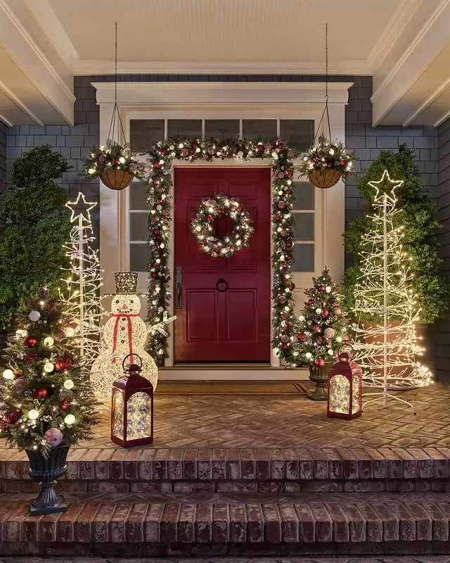 A front porch decorated for Christmas with a red door, wreaths, lit trees, string lights framing the entry door, a snowman figure, hanging plants, and lanterns on brick steps.
