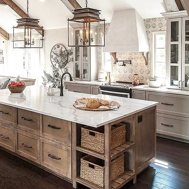 Modern French Country kitchen featuring a rustic wood island with woven baskets, marble countertops, glass lantern pendant lights, a curved plaster range hood with corbels, patterned tile backsplash, and light cabinetry for a warm farmhouse-inspired look.