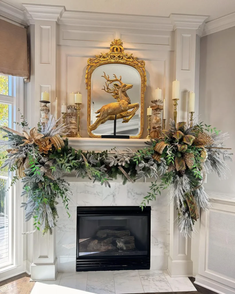 A Christmas fireplace mantel decorated with frosted greenery, gold ribbons, candles, and a gold reindeer figurine in front of a large ornate mirror.