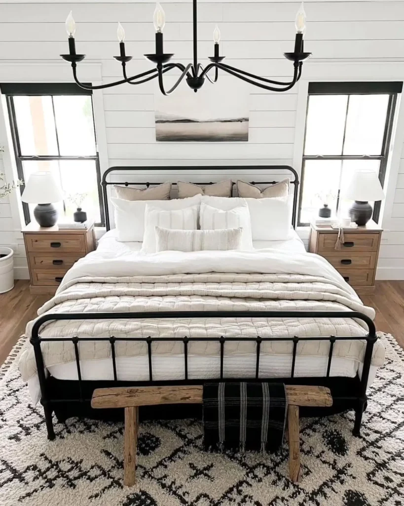 A neatly made bed with white bedding, metal frame, and a rustic bench at the foot brings industrial farmhouse bedroom charm. Two wooden nightstands with black lamps sit beside the bed, under two windows in a bright, modern space.