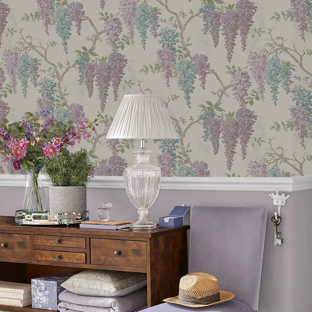 A desk with a lamp, books, and a vase of flowers is set against a wall adorned with wisteria and sand wallpaper. A light purple chair and stacked pillows are in the foreground, creating a serene workspace.