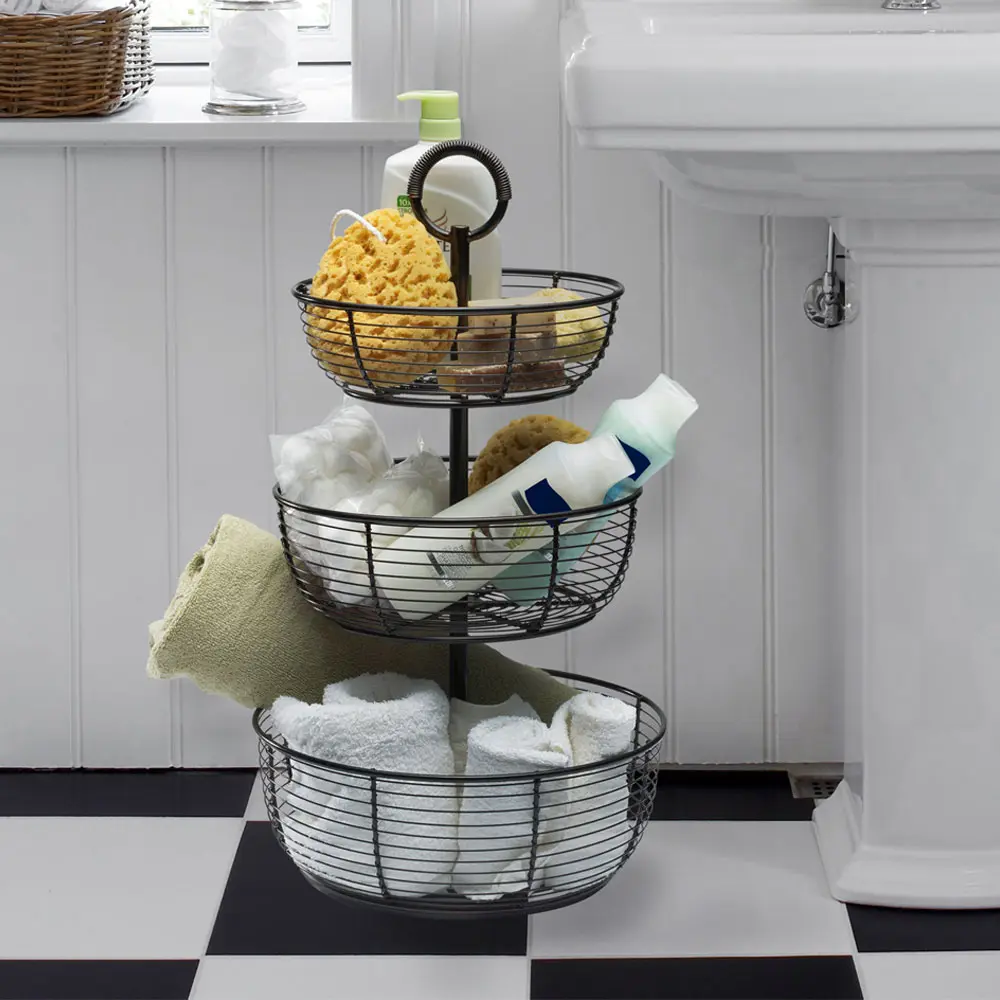3-tier wire basket bathroom organizer with towels, sponges, toiletries, and cotton balls beside pedestal sink.